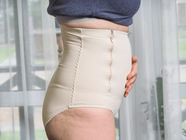 Side view of a person wearing beige high-waisted shapewear/underwear, hand resting on the abdomen near the hip.